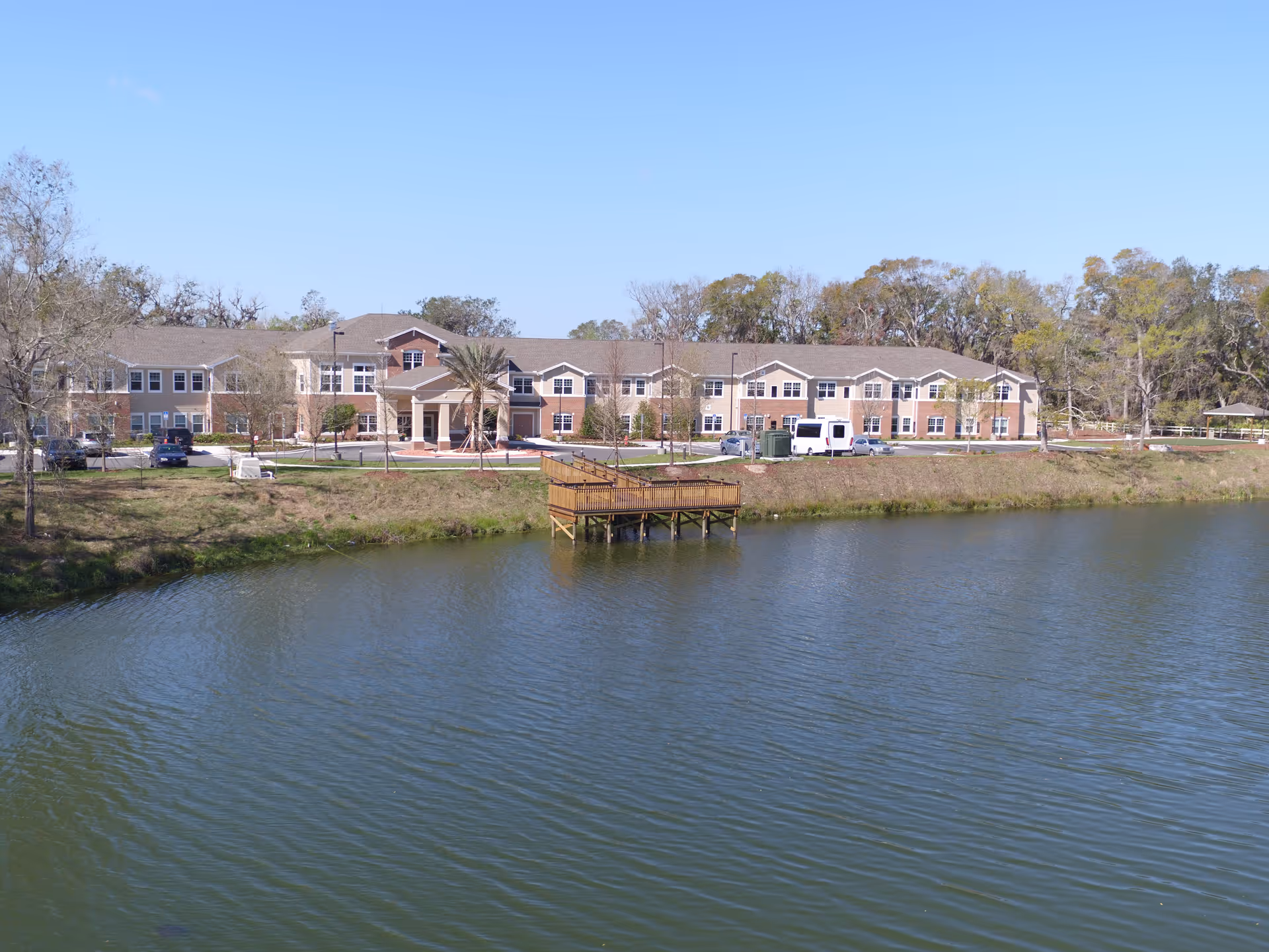 A large two-story senior living facility building named Anthem Lakes is seen across a body of water with a wooden dock extending over the water. The building has a beige and brick exterior with multiple windows and a covered entrance. There are several cars parked in front, trees surrounding the area, and a clear blue sky overhead.