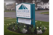 Outdoor image of a green and white sign for Montowese Center for Health & Rehabilitation, located on a grassy area with flowers planted around the base. The sign includes the text 'Montowese Health and Rehabilitation Center, Inc. Your Recovery is Our Specialty' and mentions outpatient services. Residential buildings and parked cars are visible in the background.