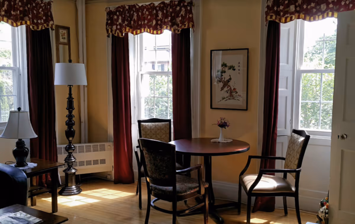 A cozy living room area with three chairs around a round wooden table, a small vase with flowers on the table, two tall windows with red curtains, a floor lamp, a table lamp on a side table, and a framed artwork on the wall.