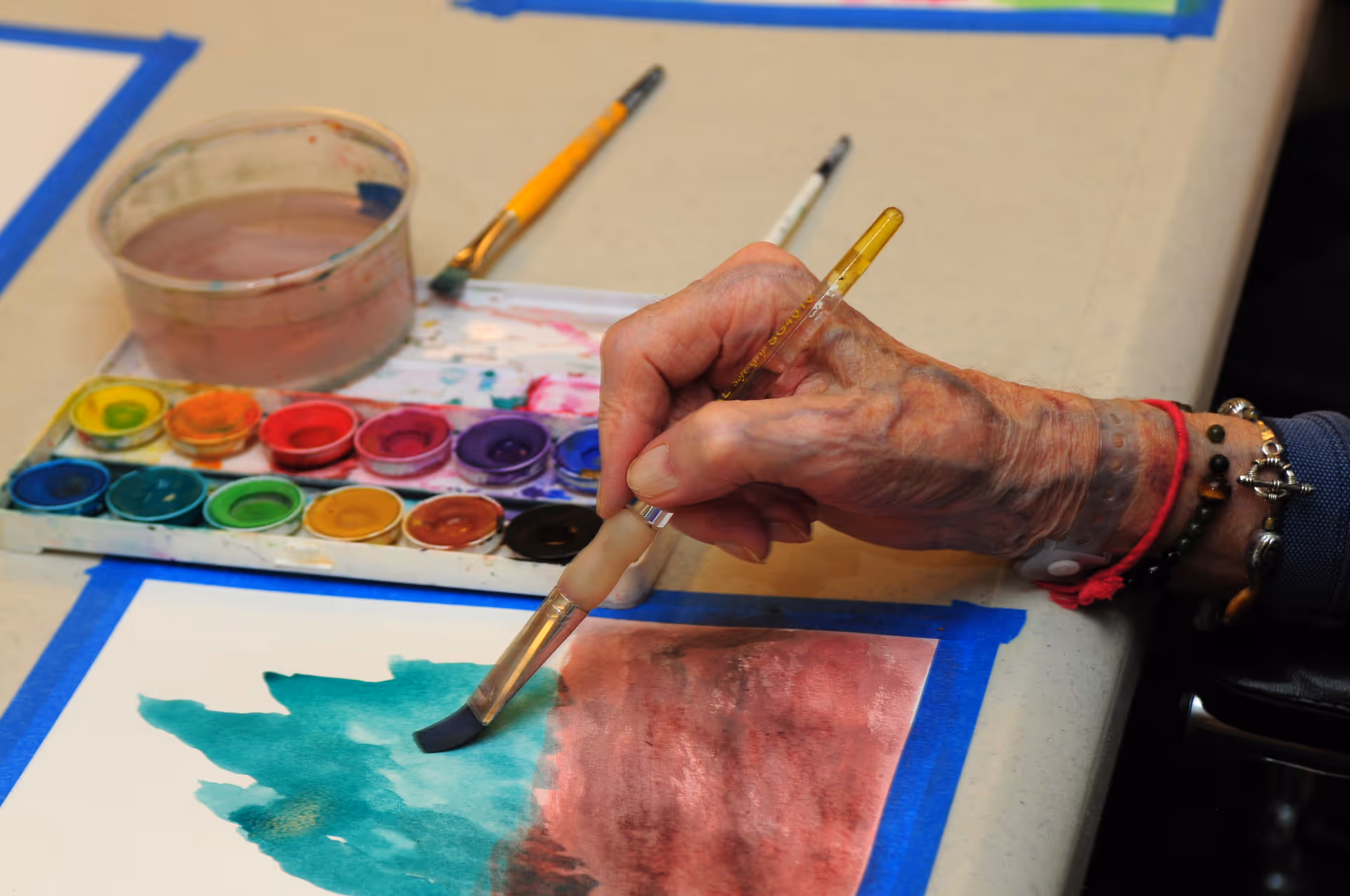 An elderly person's hand holding a paintbrush and painting with watercolors on a piece of paper taped to a table. A watercolor palette with various colors and a container of water are also visible on the table.