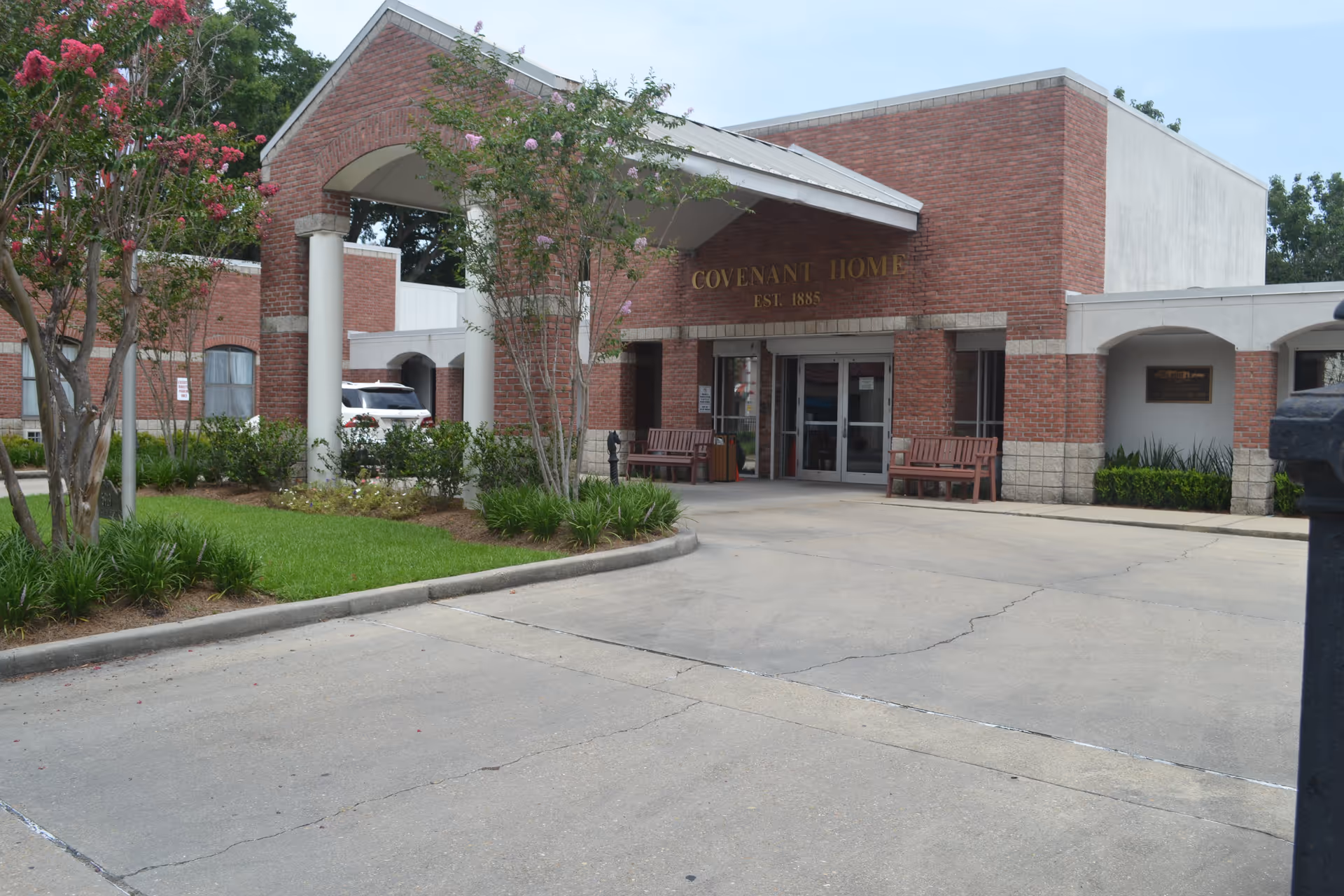 Front exterior view of Covenant Home, a brick building with a covered entrance supported by white columns, benches near the entrance, and landscaped greenery including trees and bushes.