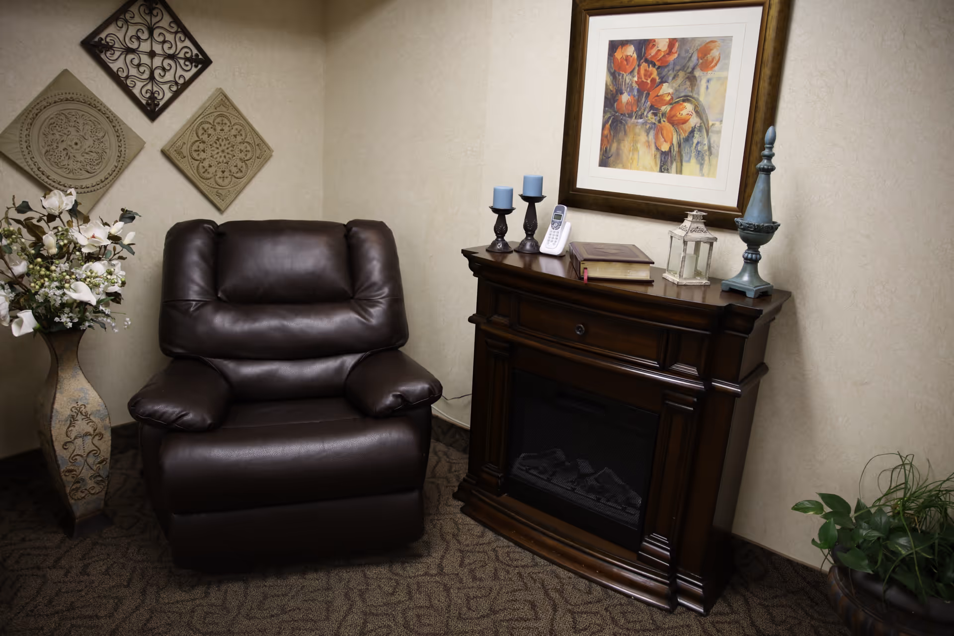 A cozy corner with a dark brown leather armchair next to a wooden electric fireplace mantel. On the mantel are decorative items including two blue candles on holders, a cordless phone, a book, a small lantern, and a tall decorative finial. Above the mantel hangs a framed painting of orange flowers. To the left of the armchair is a tall vase with white flowers, and to the right is a potted green plant. The walls are beige with three decorative wall hangings arranged diagonally.