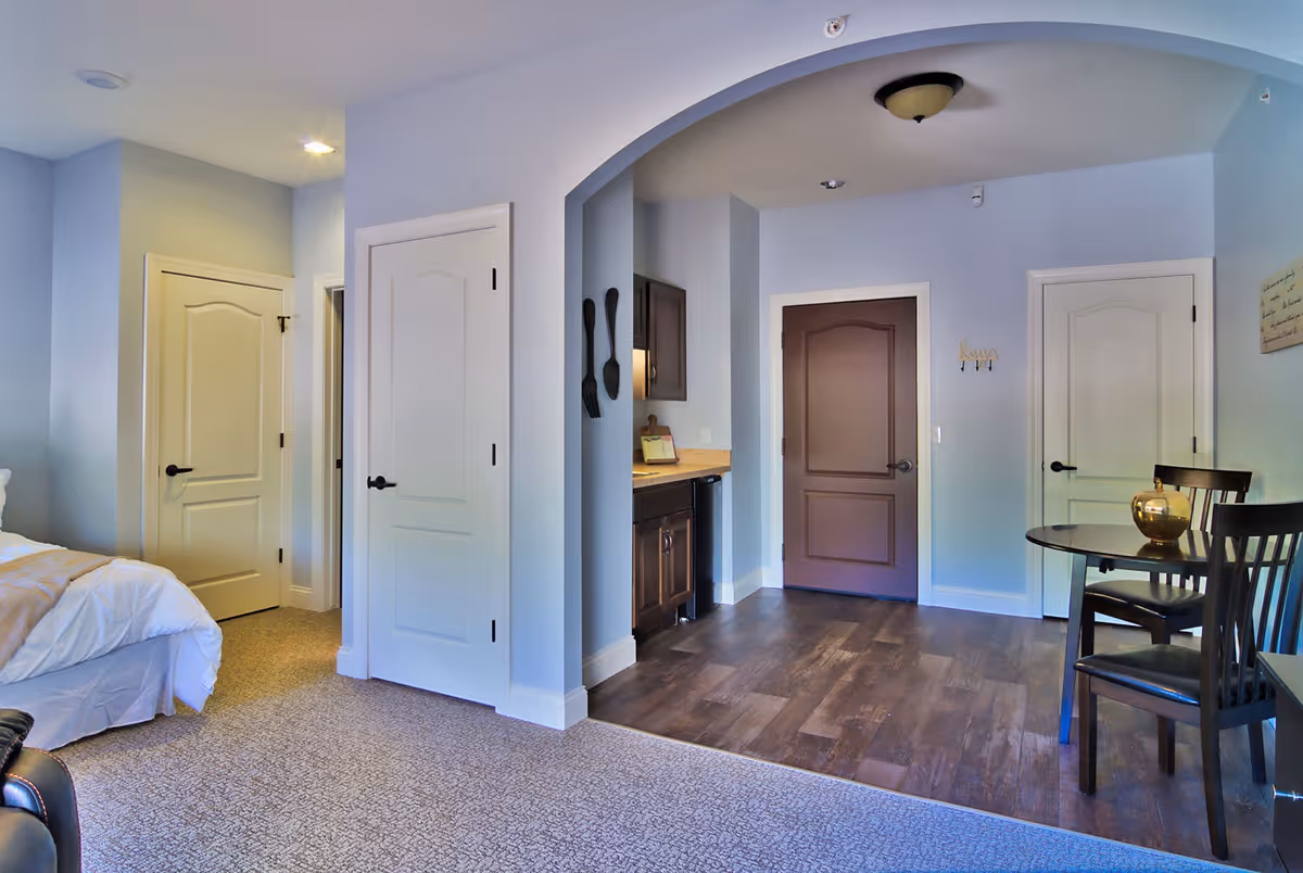 Interior view of a retirement community apartment showing a bedroom area with a bed on the left, an arched entryway leading to a small kitchenette with dark cabinets and a countertop, and a dining area with a round table and two chairs on the right. The flooring transitions from carpet in the bedroom to wood in the kitchenette and dining area. Several closed doors are visible along the walls.