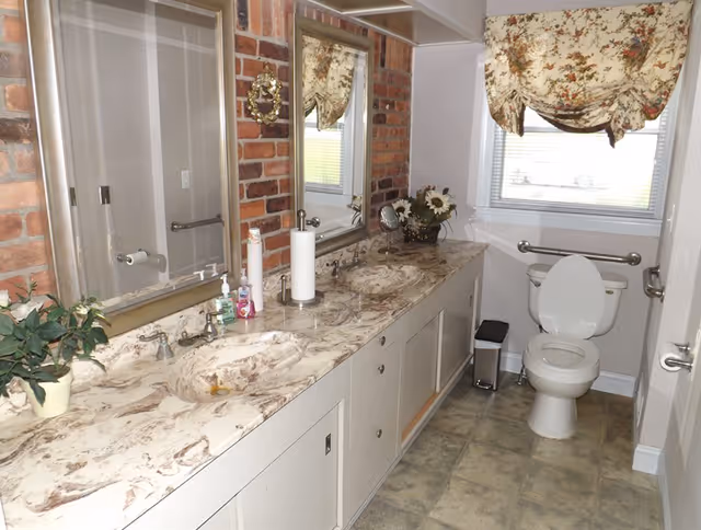 A bathroom with a long marble countertop featuring two sinks, two mirrors above the sinks, a toilet with grab bars on both sides, a window with floral curtains, a small trash can, and decorative plants and flowers on the countertop.