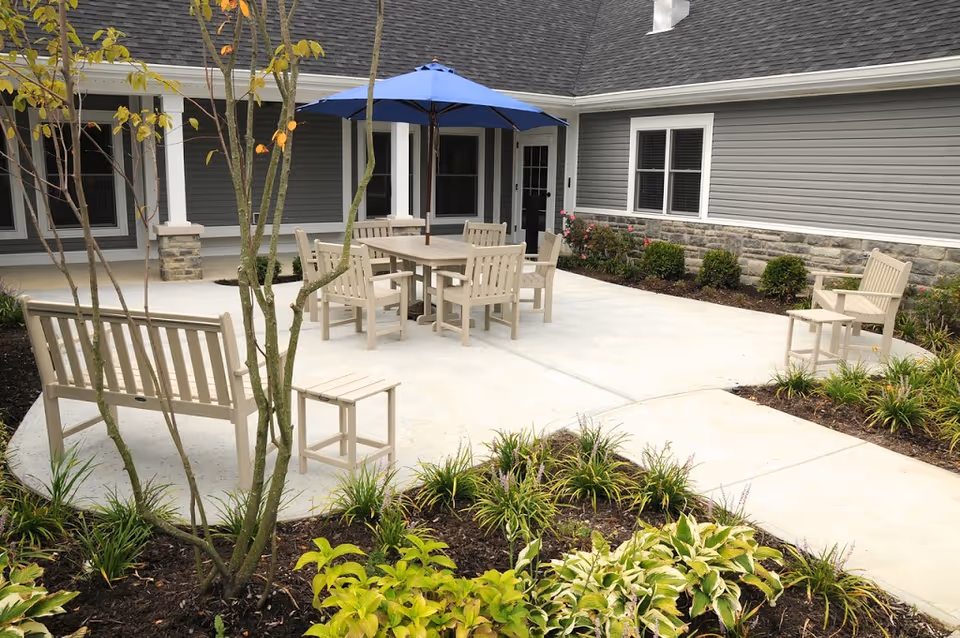 Outdoor courtyard with beige chairs and benches around a table under a blue umbrella on a paved patio, surrounded by landscaping in front of a gray-sided building.