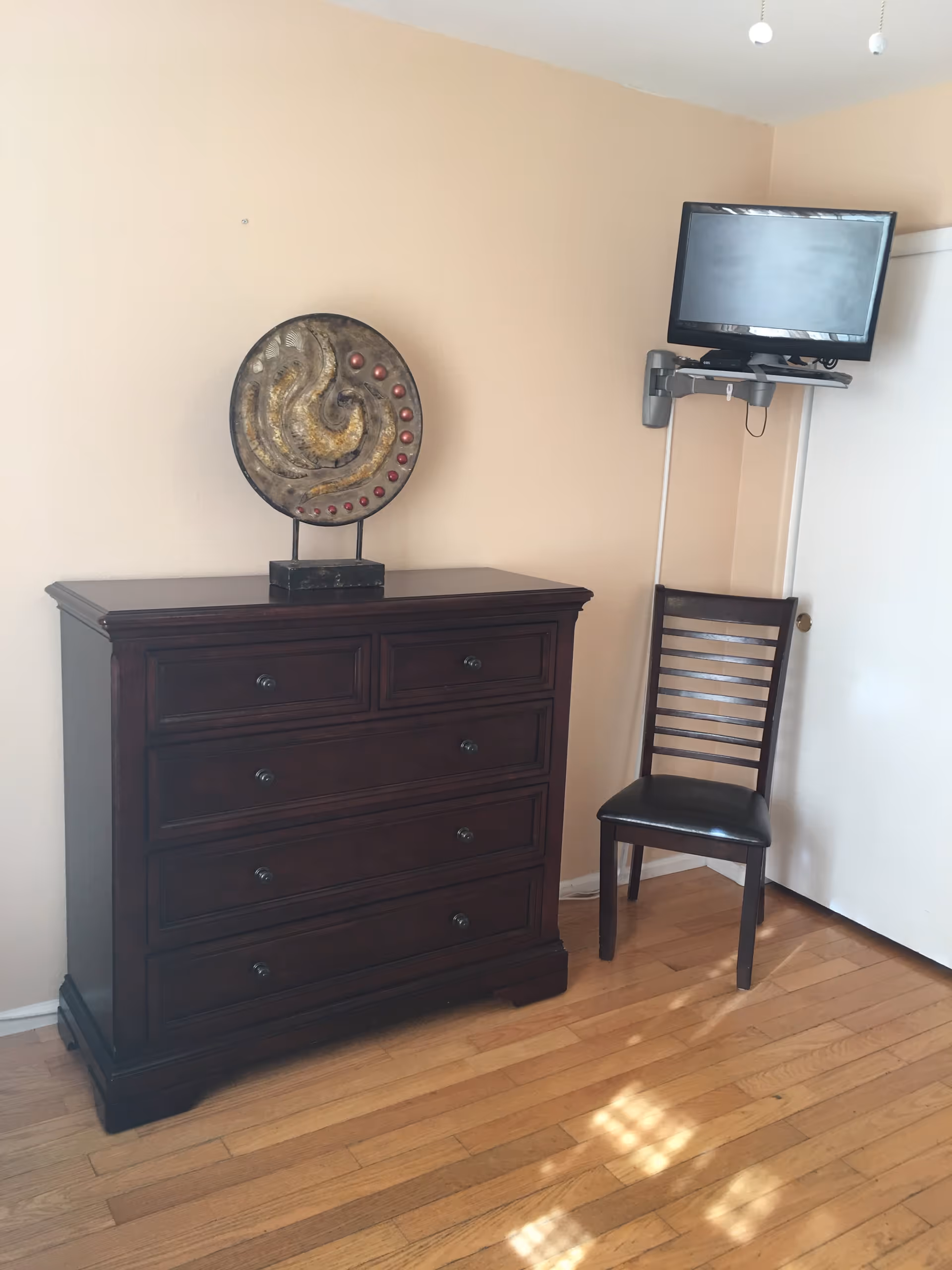 A corner of a room with a dark wooden dresser topped with a decorative circular sculpture, a wooden chair with a black cushioned seat, a wall-mounted flat screen TV, and light-colored walls and hardwood flooring.