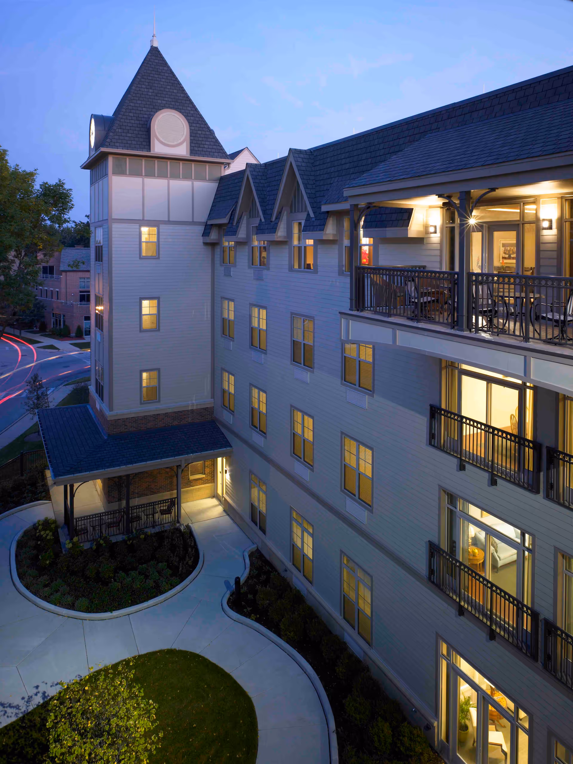 Exterior view of a multi-story senior living facility building at dusk with illuminated windows and a covered entrance. The building features a tower with a steep roof and balconies with outdoor seating. A curved walkway and landscaped greenery are visible in the foreground.