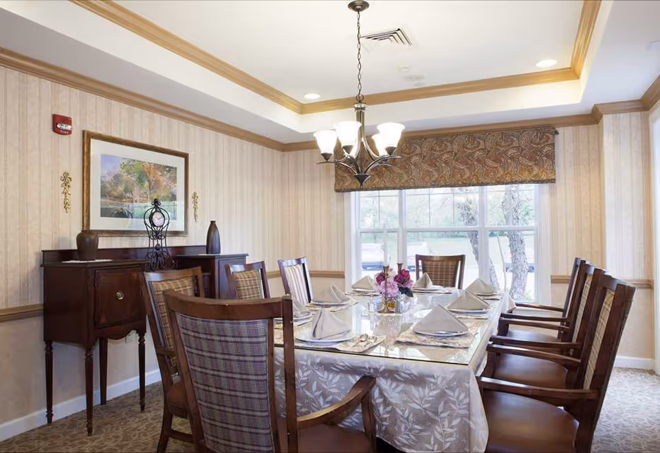 Formal dining room with a long set table for eight, wooden chairs, a chandelier, sideboard and large window.