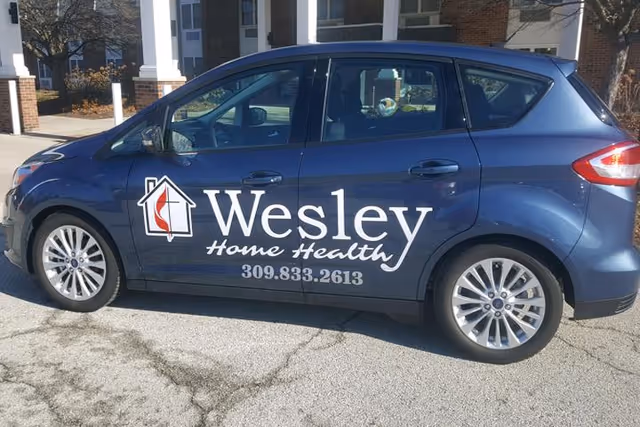 A blue car parked outside a building with the logo and text 'Wesley Home Health 309.833.2613' displayed on the side of the vehicle.