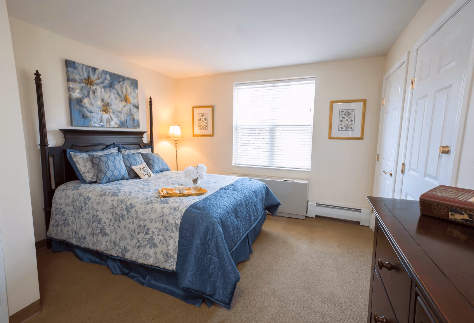 A neatly made bed with blue and white floral bedding in a bedroom. The bed has multiple pillows and a wooden headboard. A tray with a flower vase and cups is placed on the bed. There is a window with blinds letting in natural light, two framed pictures on the walls, a floor lamp in the corner, and a wooden dresser with books on top.