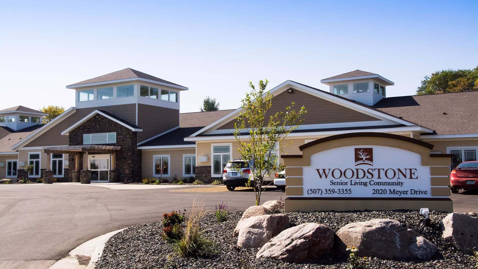 Exterior view of Woodstone Senior Living Community building with a clear blue sky, a paved driveway, landscaped rocks and plants, and a sign displaying the facility name, phone number, and address.