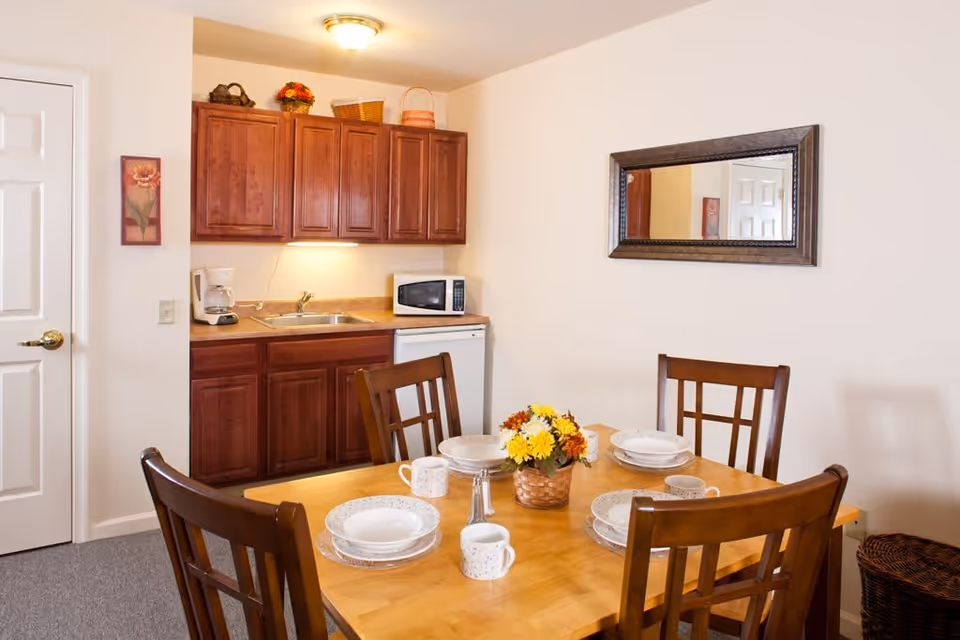 A small dining area with a wooden table set for four, including plates, bowls, mugs, and a centerpiece with yellow and orange flowers. Behind the table is a kitchenette with wooden cabinets, a sink, a coffee maker, a microwave, and a small refrigerator. A rectangular mirror hangs on the wall above the dining table, and a door is visible to the left.