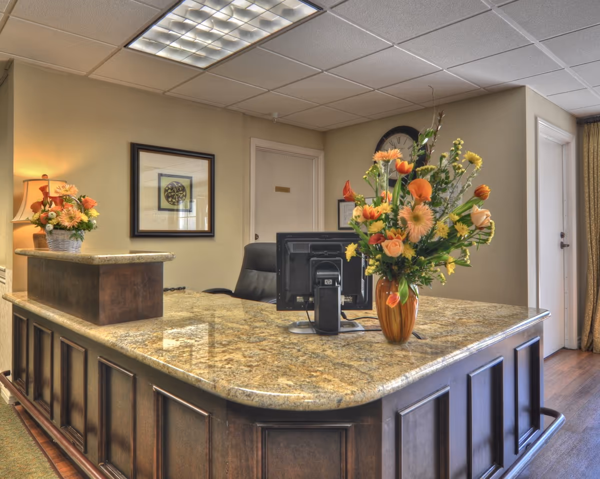 Reception desk area with a granite countertop and dark wood paneling. There is a computer monitor on the desk, a large vase with orange and yellow flowers, a lamp, and a framed picture on the beige wall behind the desk. A clock is mounted on the wall above a door in the background.