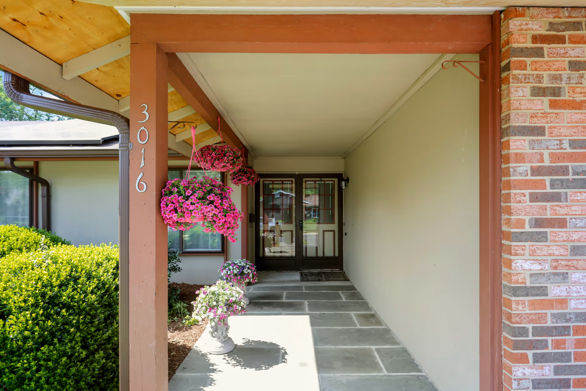 Covered entrance walkway to a building with a brown wooden post marked with the number 3016. Hanging baskets with pink flowers and a white flower pot with purple and white flowers are placed along the walkway. The entrance door is glass with a brown frame, and there is a brick wall on the right side.