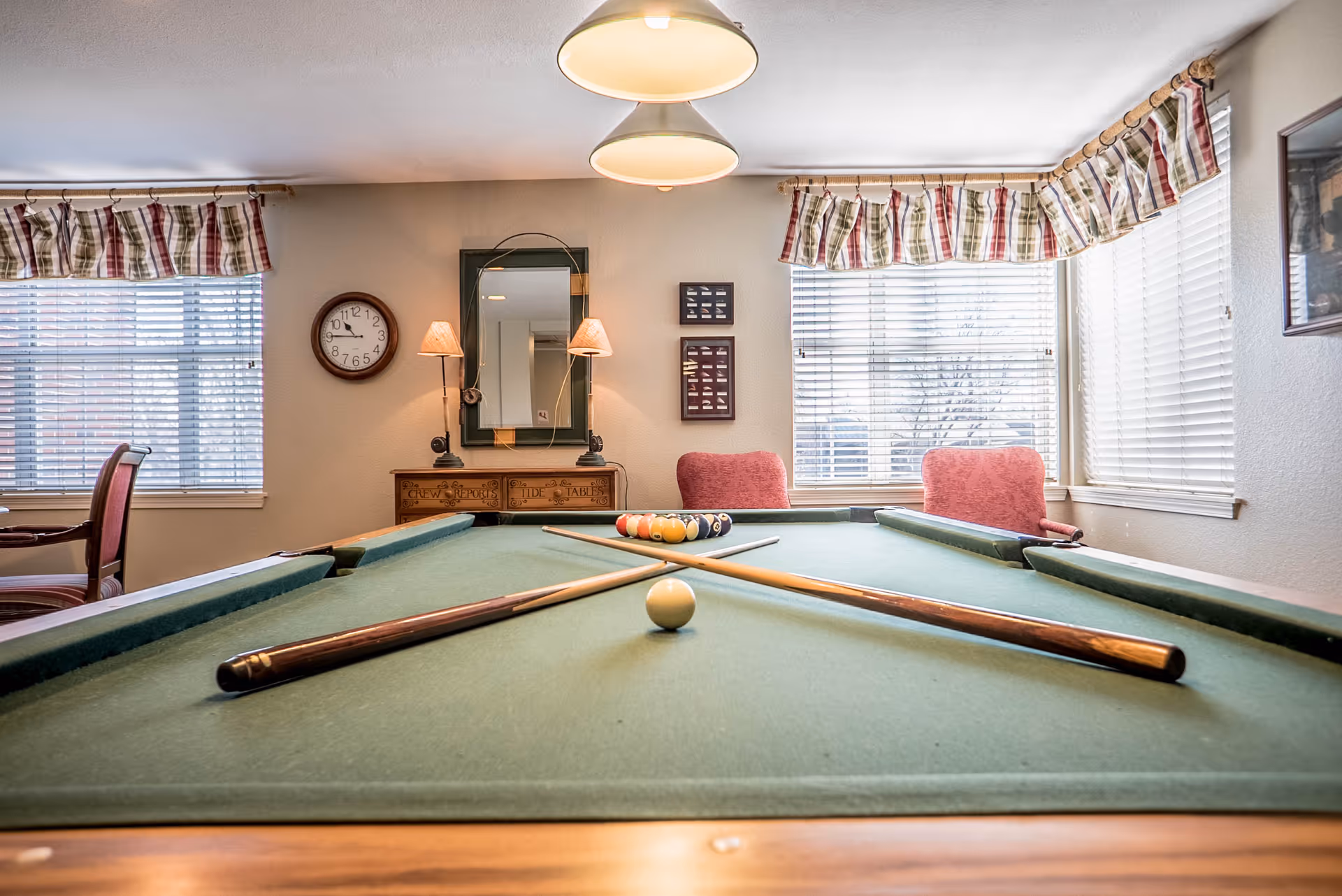 A pool table with two crossed cues and a cue ball in the foreground, set in a well-lit room with large windows covered by plaid valances. The room also features a wall clock, a mirror with two wall lamps, framed pictures, and two red upholstered chairs near the windows.