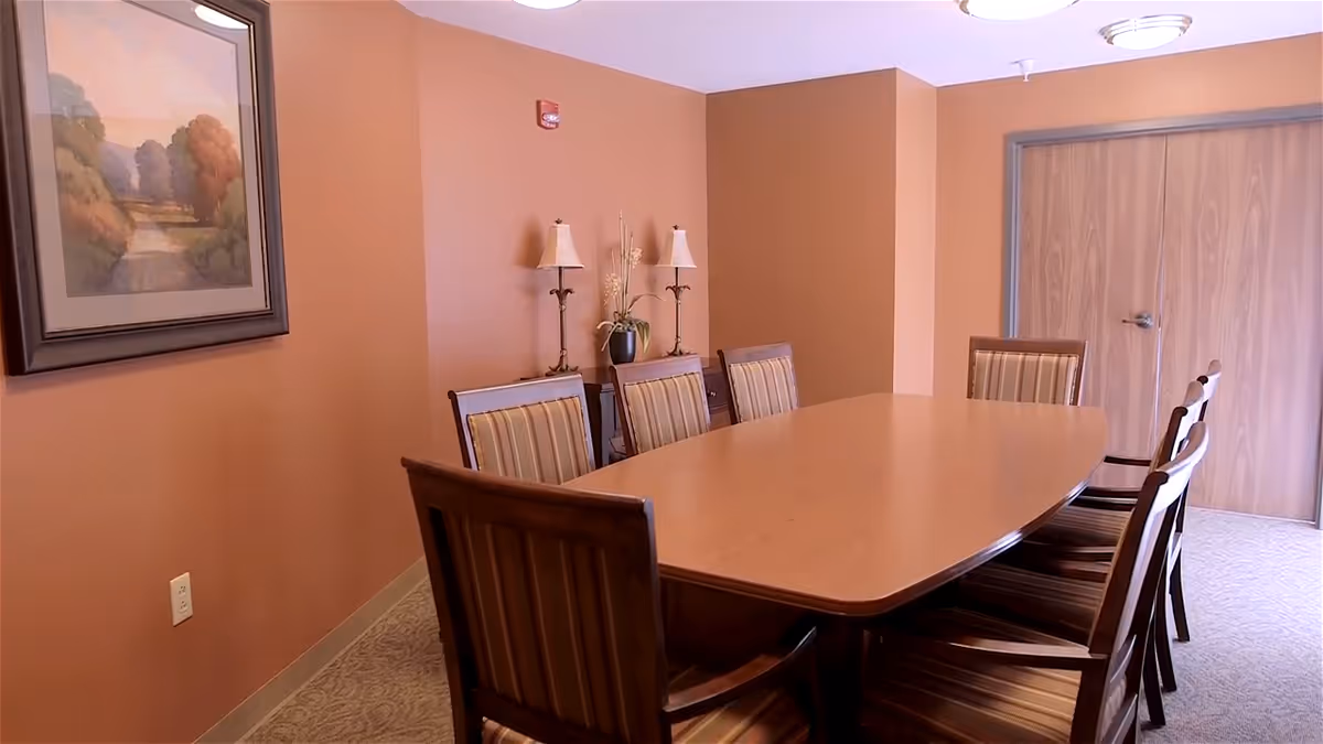 A dining room with a long wooden table surrounded by eight wooden chairs with striped cushions. The walls are painted a warm brown color, and there is a framed landscape painting on the left wall. Two table lamps and a vase with flowers are placed on a sideboard against the back wall. Double wooden doors are visible at the far end of the room.