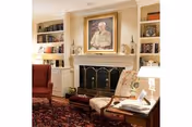 A cozy living room with a fireplace in the center, flanked by built-in bookshelves filled with books and decorative items. Above the fireplace is a framed portrait of an elderly man. In front of the fireplace is a cushioned armchair with a floral pattern, a small wooden side table with a lamp and books, and a red upholstered chair. The room features a patterned rug on the floor and soft lighting from table lamps.
