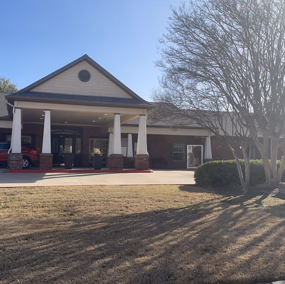 Front entrance of a single-story senior living building with a covered porte-cochère, white columns, driveway and trees on the lawn.