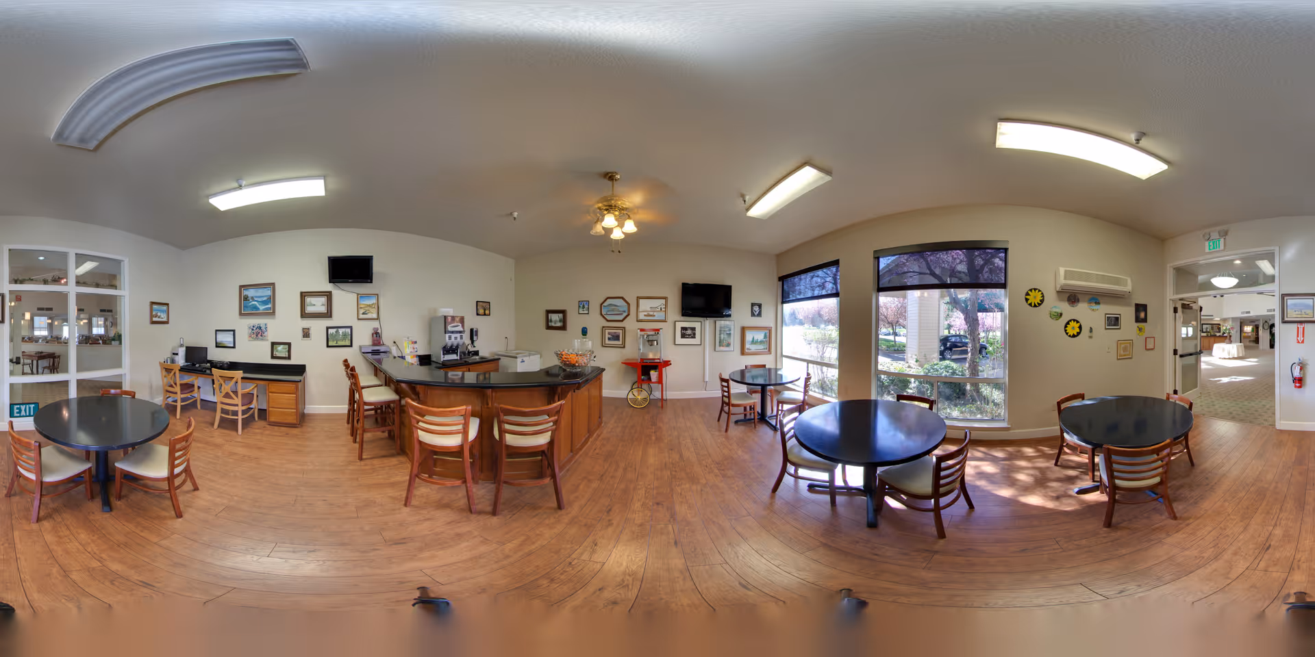 A bright and spacious common dining area with several round tables and wooden chairs arranged on a wooden floor. The room features large windows letting in natural light and offering views of outdoor greenery. There is a central counter with bar stools, a popcorn machine, and two wall-mounted televisions. The walls are decorated with various framed pictures and artwork. Ceiling lights and a ceiling fan provide additional lighting.