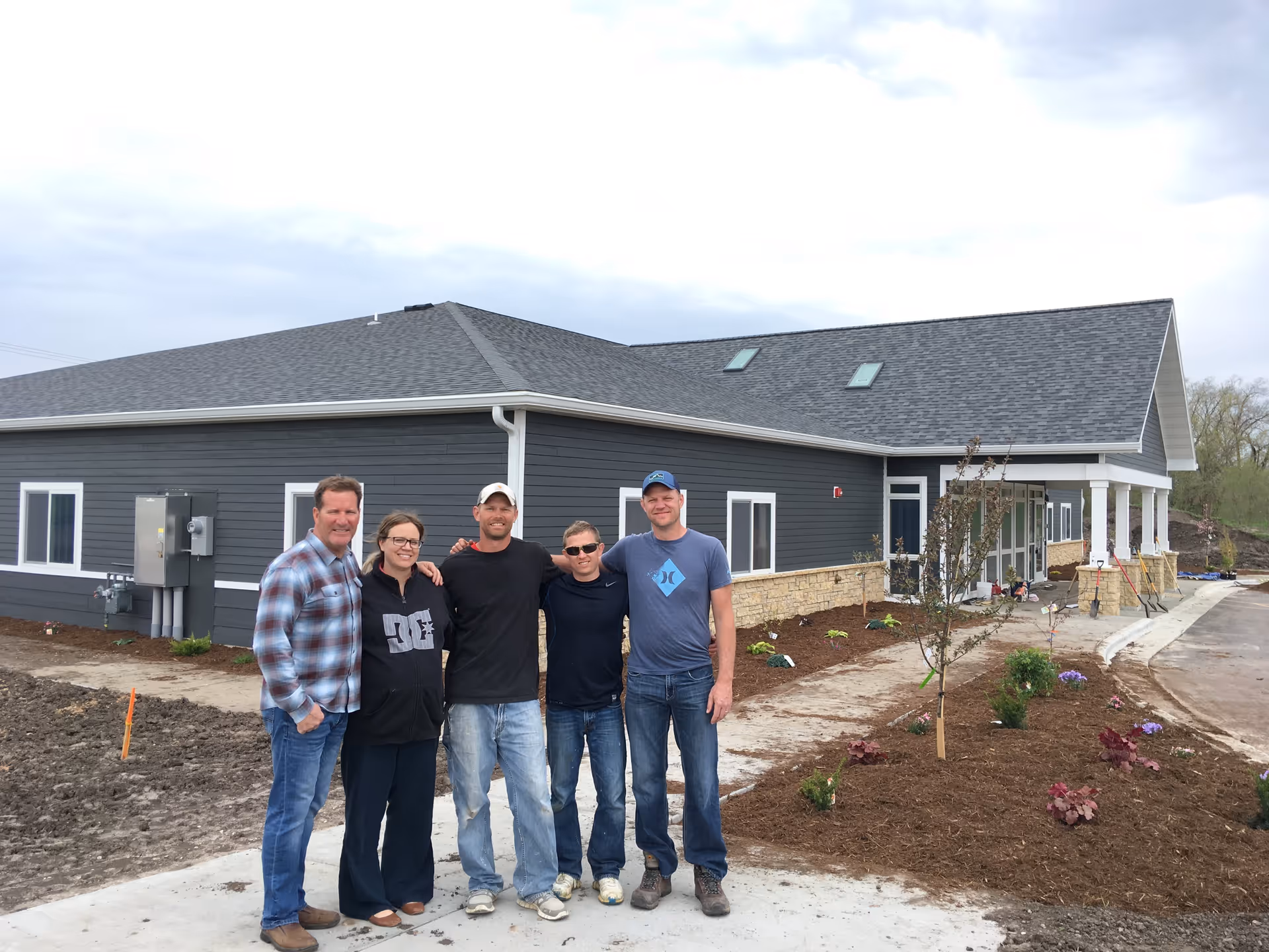 Five people standing outside in front of a newly constructed single-story building with gray siding and a stone base. The area around the building is landscaped with small plants and a young tree, and the sky is overcast.