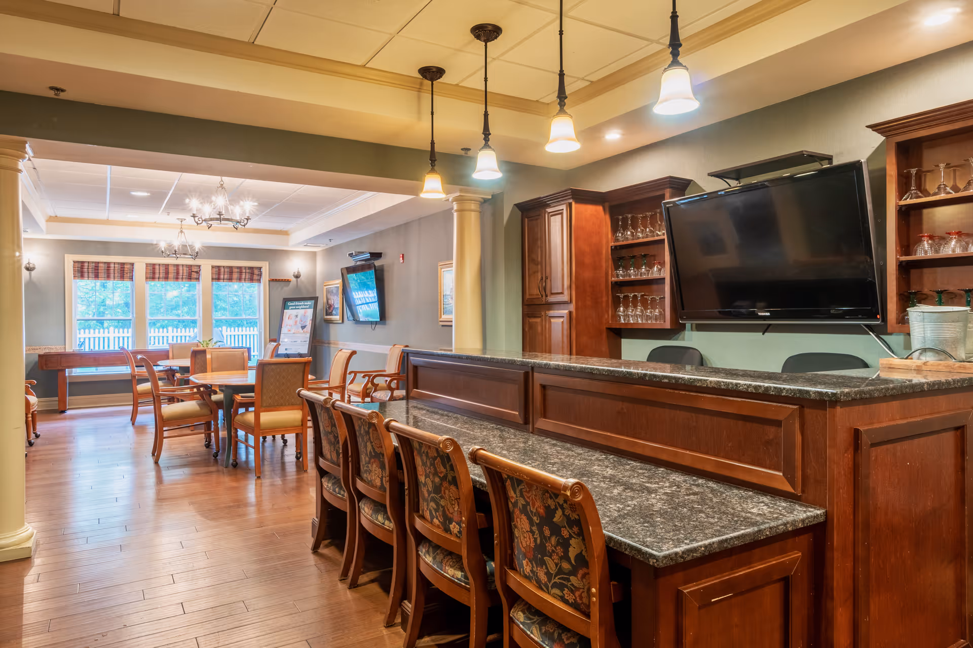 Interior view of a senior living facility common area with a long granite countertop bar and wooden chairs in the foreground. Behind the bar are wooden cabinets with glassware and a mounted flat-screen TV. In the background, there is a dining area with multiple tables and chairs, large windows with plaid valances, and chandeliers hanging from the ceiling.