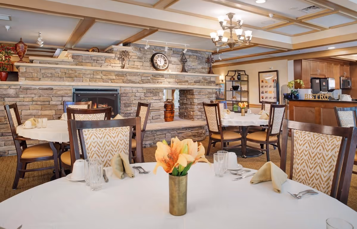 Bright, elegant dining room with round tables set for meals, a stone fireplace wall and a bistro counter in the background.