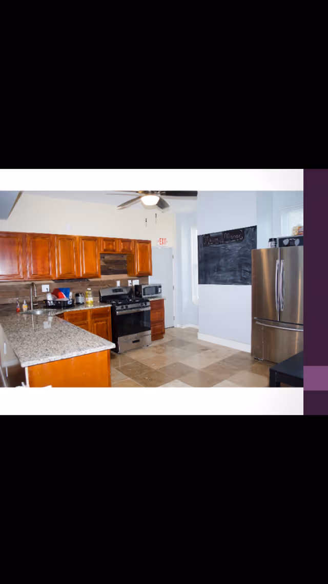 A kitchen with wooden cabinets, granite countertops, a stainless steel refrigerator, a gas stove, a microwave, and a ceiling fan with light. There is a black chalkboard on the wall and beige tiled flooring.