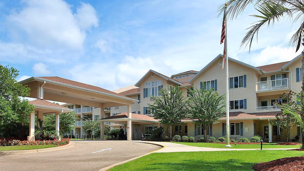 Exterior view of Cherry Laurel - Sky Active Living facility showing a large, multi-story building with beige walls and red roofs, surrounded by well-maintained landscaping including green grass, trees, and flower beds. There is a covered driveway entrance and an American flag on a flagpole in front of the building under a partly cloudy sky.