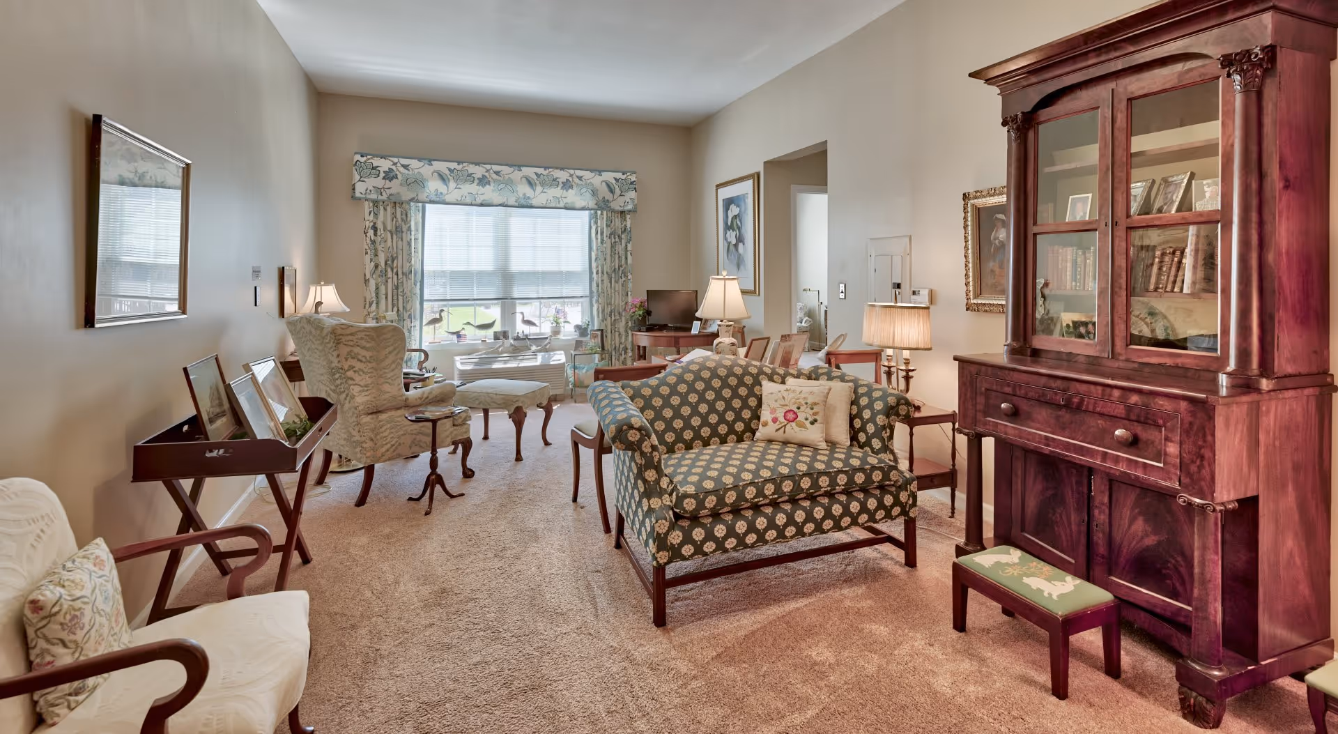 Cozy sunlit living room with patterned loveseat, armchairs, side tables, lamps, and a large wooden display cabinet.