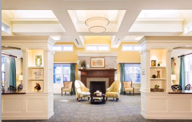 A bright and spacious living room area with a central fireplace, two yellow patterned armchairs facing each other with a small round coffee table between them. The room features large windows with teal curtains, built-in shelves with decorative items, and a coffered ceiling with a modern chandelier.