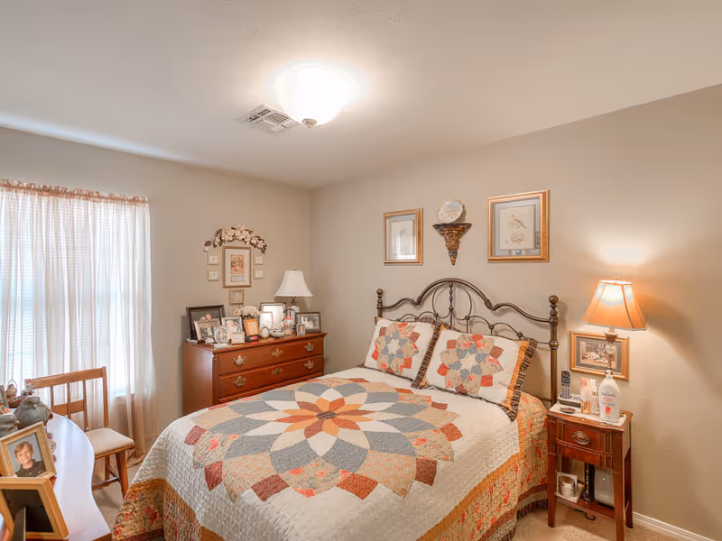 A cozy bedroom with a bed featuring a colorful quilt and matching pillows. The room has a wooden dresser with framed photos and a lamp, a nightstand with a lamp and personal items, and a window with sheer curtains allowing natural light to enter.