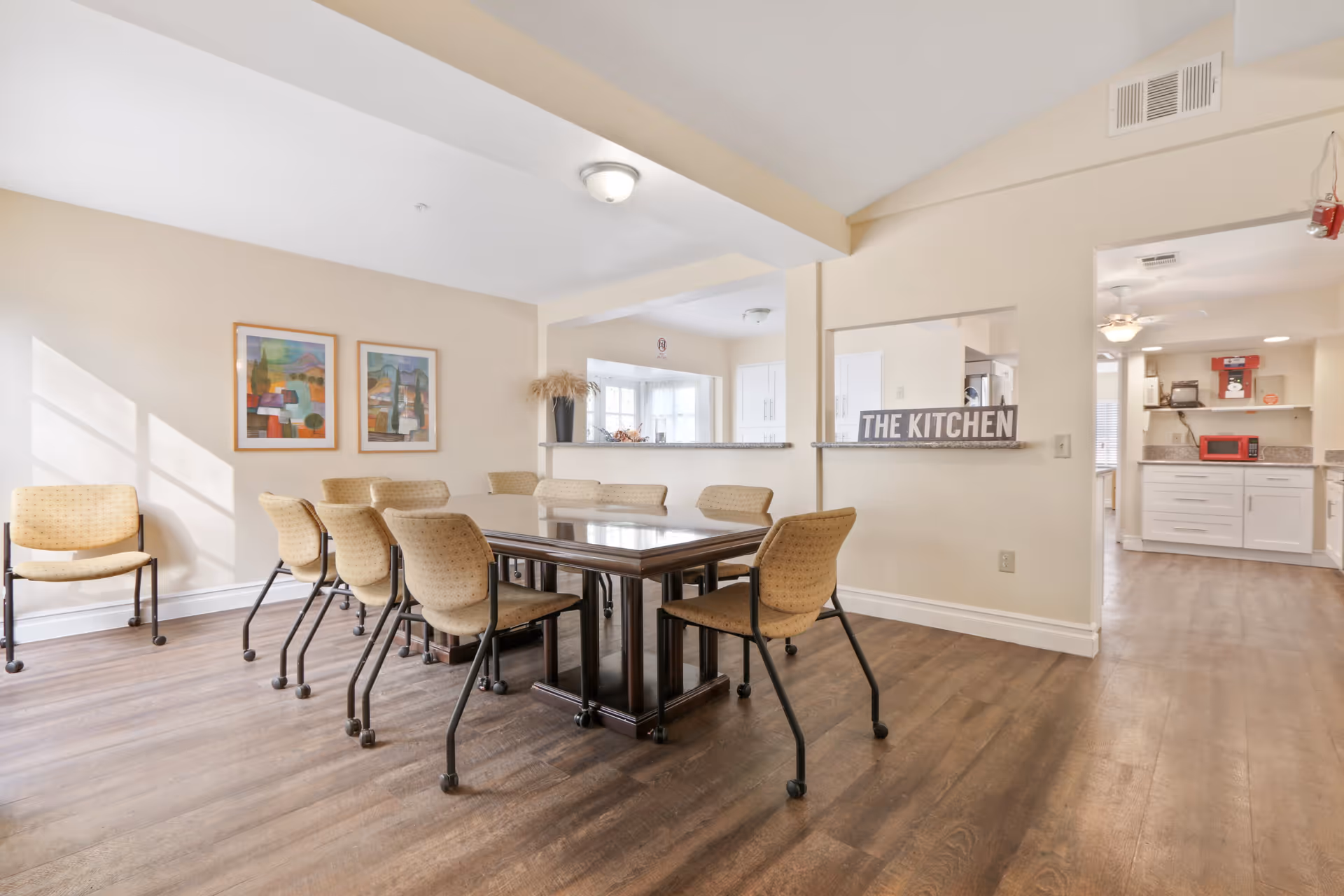 Bright dining area with a rectangular table surrounded by beige upholstered chairs and a pass-through window labeled "THE KITCHEN" leading to the adjacent kitchen.