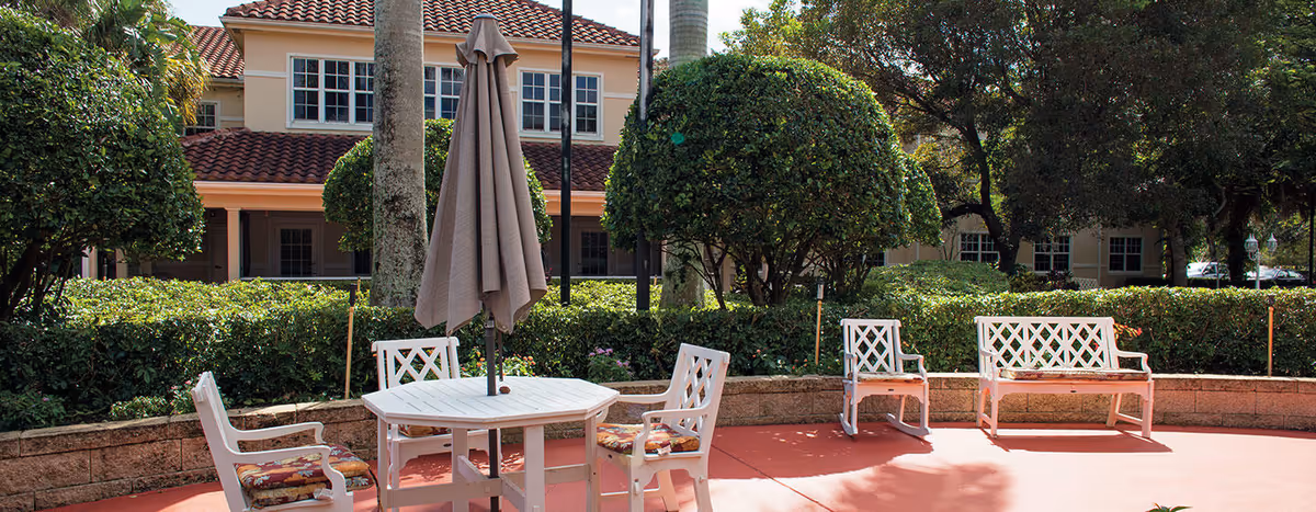 Outdoor patio with white chairs, a table and umbrella in front of a two-story building and landscaped hedges.