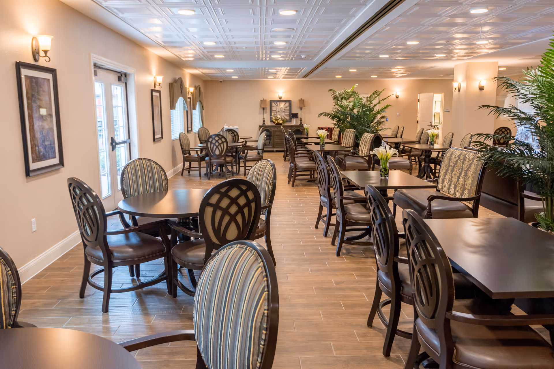 A spacious dining room with multiple dark wood tables and chairs arranged neatly. The room features wood flooring, beige walls, framed artwork, wall sconces for lighting, and large windows with blinds. There are also several green plants placed around the room, creating a welcoming atmosphere.