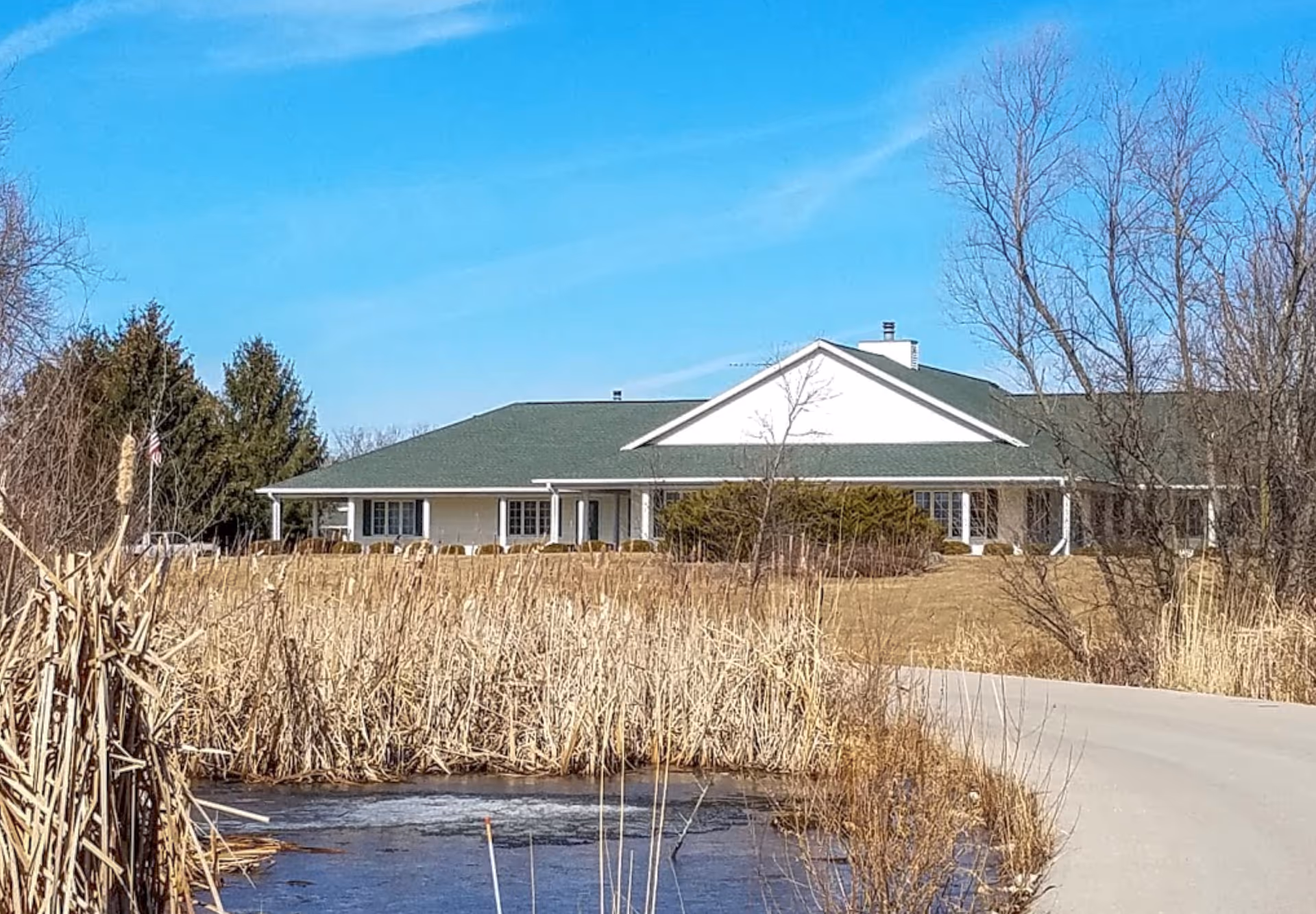 A single-story building with a green roof and white exterior walls, surrounded by dry tall grass and leafless trees under a clear blue sky. A paved driveway curves in front of the building, and there is a small pond with some ice in the foreground.