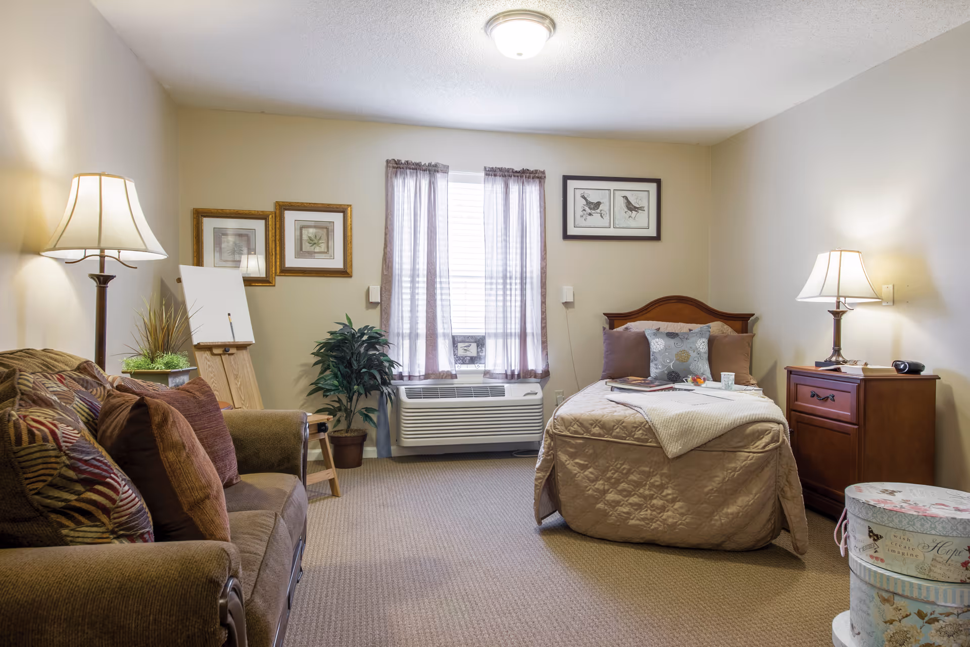A cozy senior studio room with a bed, sofa, side tables and lamps, a window with an air conditioner, and decorative artwork.