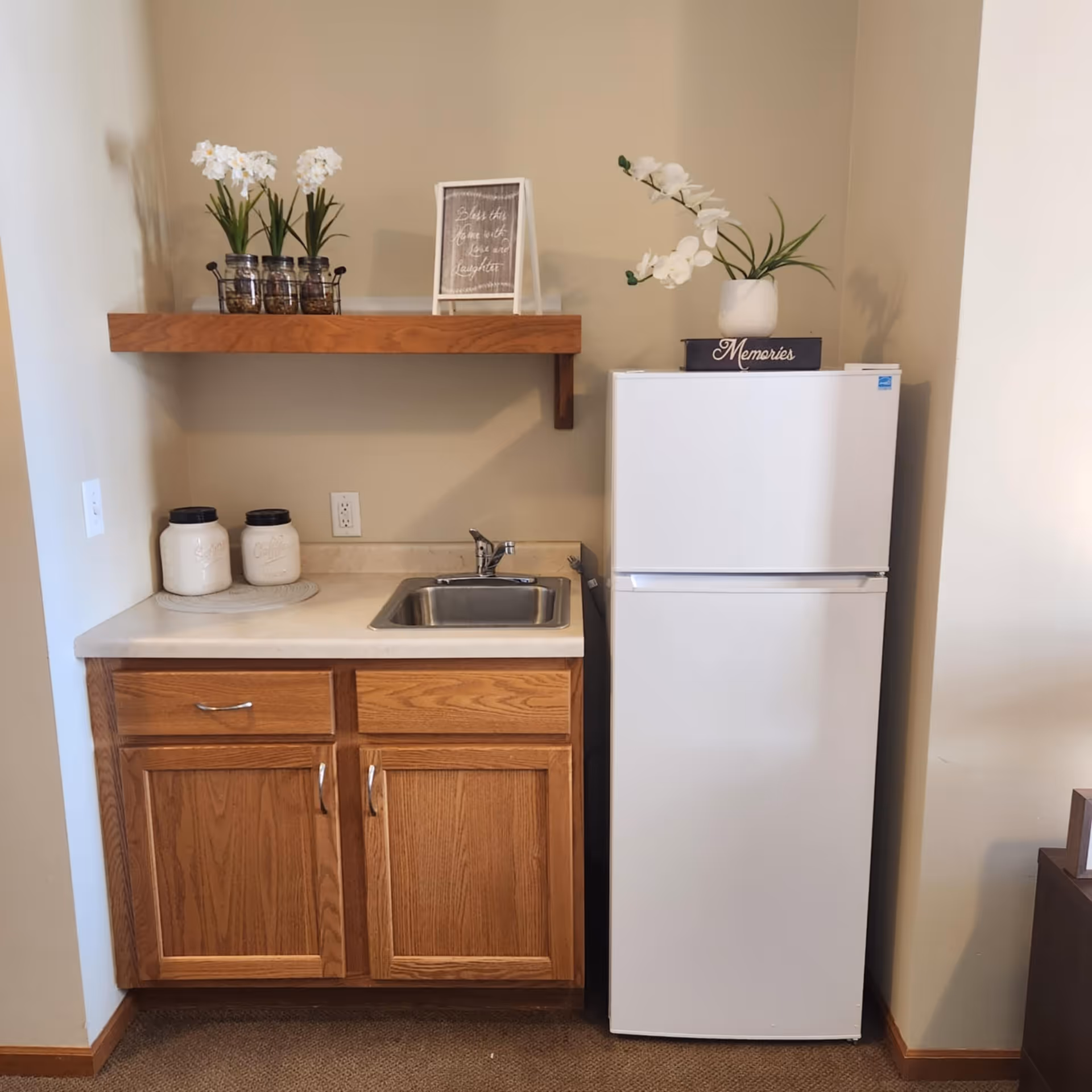 A small kitchenette area with a white refrigerator on the right and a wooden cabinet with a countertop and a small sink on the left. Above the countertop is a wooden shelf holding three small potted plants and a framed sign. On top of the refrigerator is a white vase with white flowers placed on a black box labeled 'Memories'.