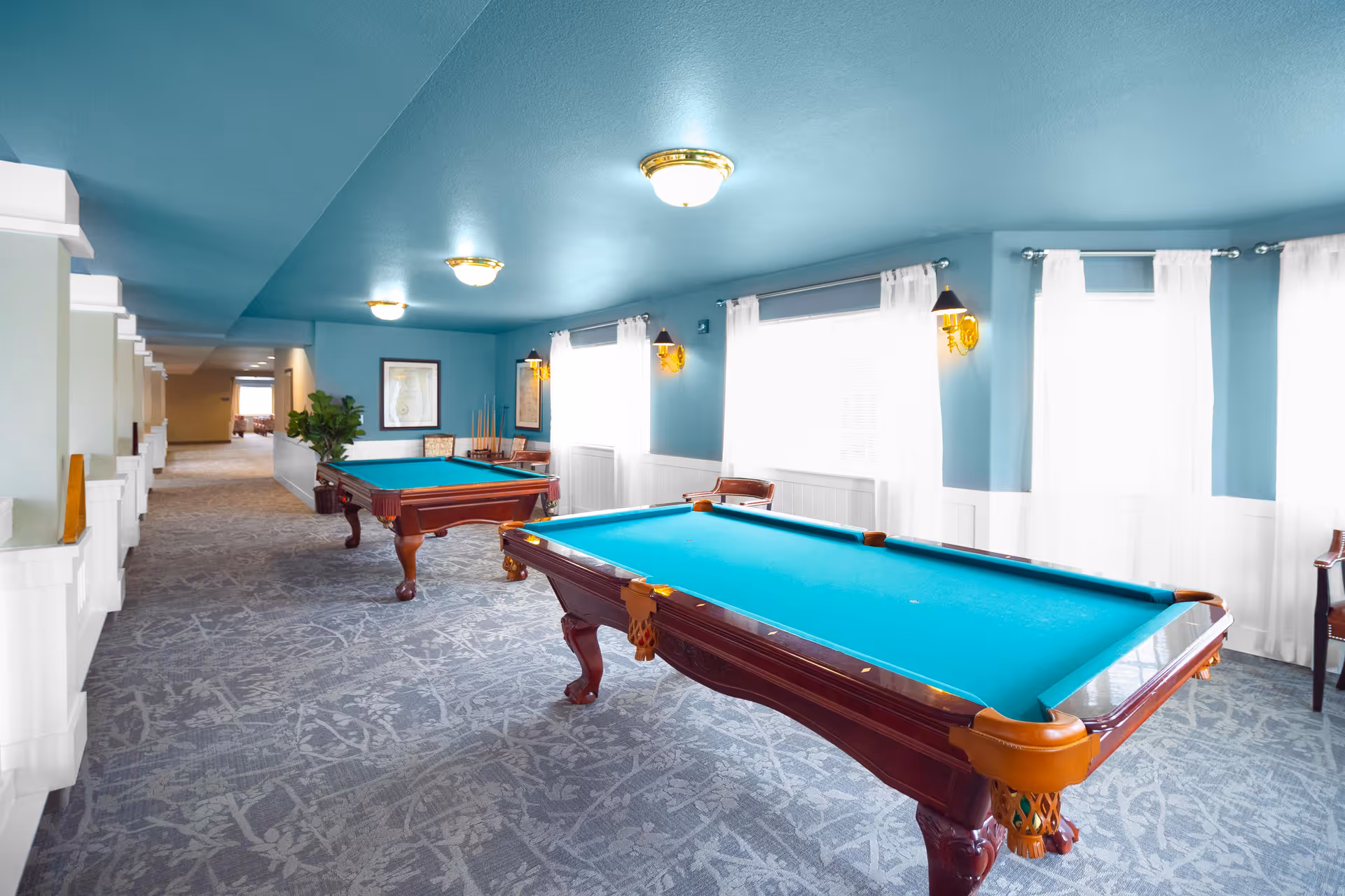 A bright recreational room with two pool tables featuring teal felt tops. The room has light blue walls and ceiling, large windows with white curtains allowing natural light to fill the space, patterned carpet flooring, and several chairs along the walls. There are framed pictures on the walls and ceiling lights providing additional illumination.
