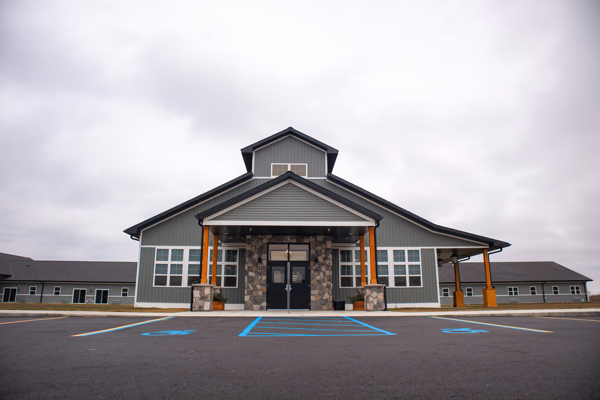 Front exterior view of a large single-story building with gray siding, stone accents around the entrance, and wooden pillars supporting a covered porch. The building has multiple windows and a parking lot with handicap spaces in front. The sky is overcast.