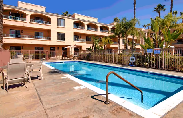 Outdoor swimming pool area at Las Villas Del Norte with clear blue water, surrounded by a concrete deck with chairs and tables. The pool is fenced, and there are palm trees and a three-story building with balconies in the background under a clear blue sky.