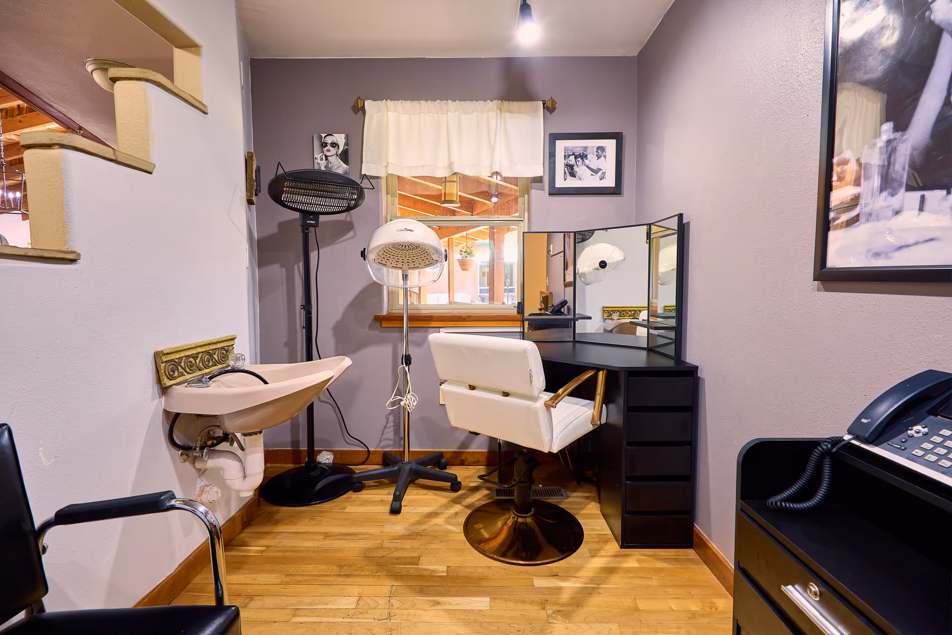 A small salon or beauty station area with a white salon chair in front of a black vanity with a three-panel mirror. There is a hair dryer hood on a stand, a wall-mounted sink, a black chair, and a telephone on a black cabinet. The walls are painted light purple, and there are framed black and white photos on the walls. A window with a white curtain is above the vanity.