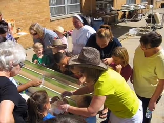 A group of children and adults gathered around a long water table outdoors, engaging in an activity. The setting appears to be a sunny courtyard with a brick building in the background. One adult wearing a hat and bright yellow shirt is assisting the children, while others watch and participate.