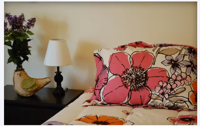 Close-up view of a bed with floral-patterned bedding in shades of pink, orange, and purple. Next to the bed is a dark wooden nightstand with a small white lampshade on a black lamp base and a decorative bird figurine with a small bouquet of purple flowers.