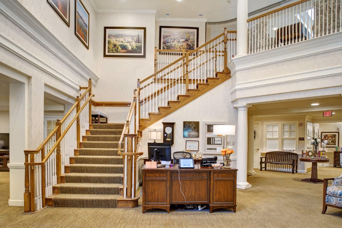 Interior view of a senior living facility lobby with a carpeted staircase featuring wooden handrails and white balusters. A wooden reception desk with a computer, lamp, and decorative items is positioned in front of the stairs. The walls are adorned with framed artwork, and there are columns and seating areas visible in the background.