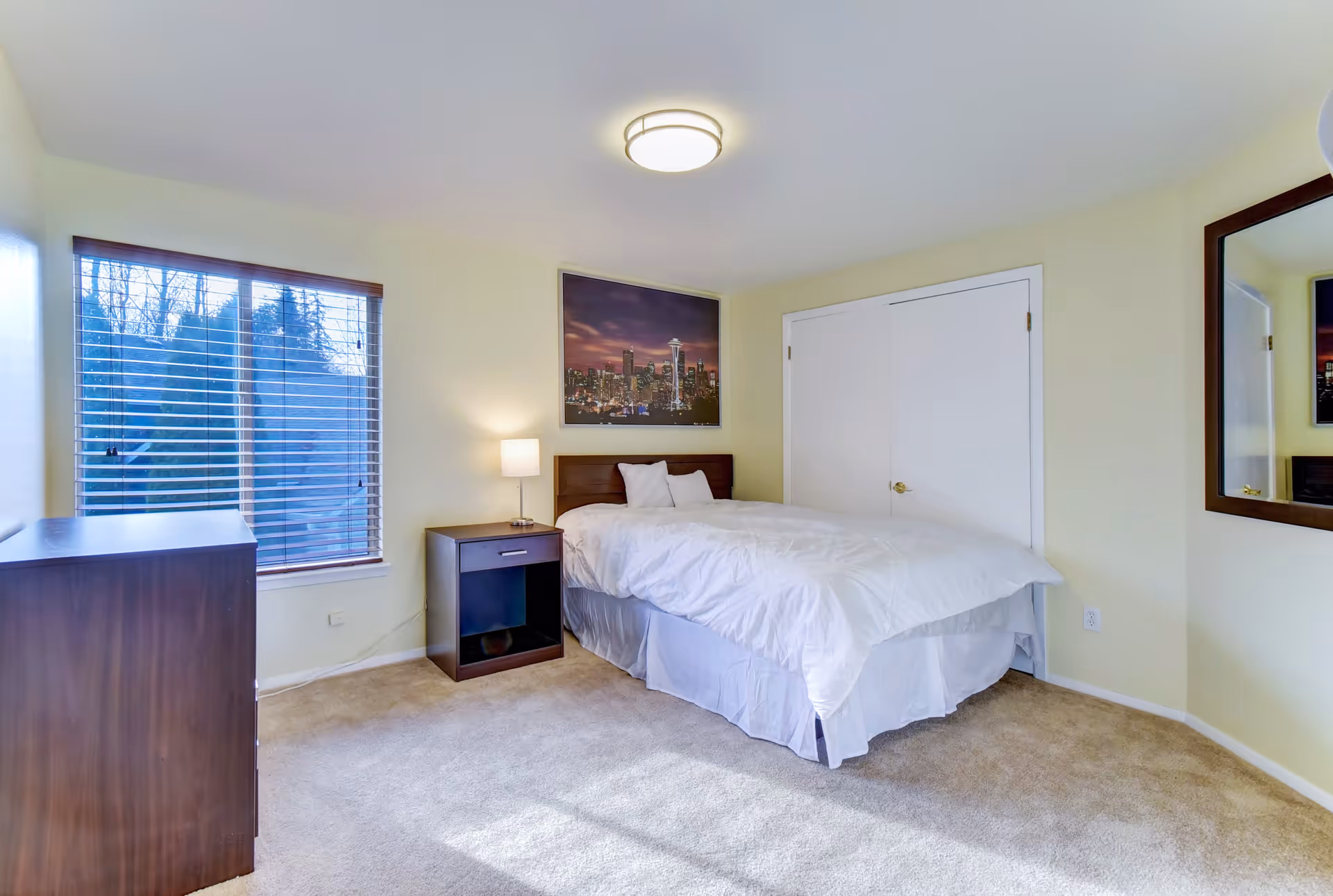 A bedroom with a bed covered in white bedding, a wooden nightstand with a lamp, a wooden dresser, a window with blinds, a large mirror on the wall, and a framed picture of a city skyline above the bed.