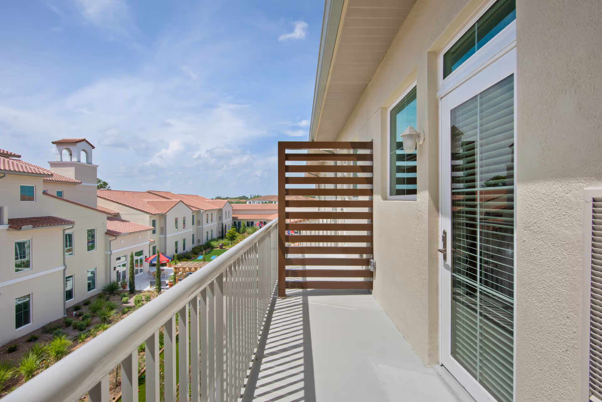 A balcony with a white railing and a wooden privacy screen at the end. The balcony is attached to a beige building with a glass door and windows with white blinds. In the background, there are multiple beige buildings with red tile roofs under a partly cloudy blue sky.