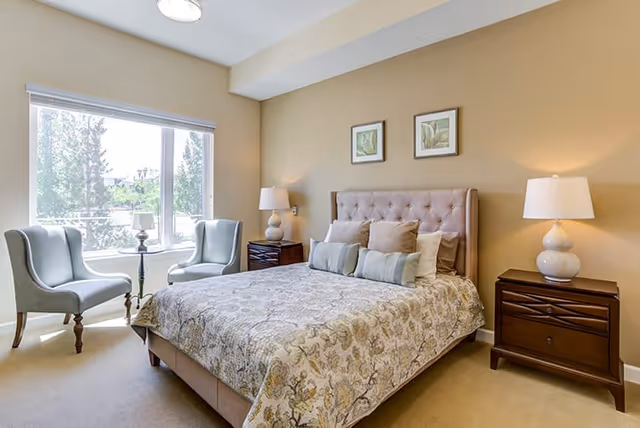 A well-lit bedroom with a large window showing trees outside. The room features a bed with a patterned bedspread and multiple pillows, a tufted headboard, two wooden nightstands each with a white lamp, and two light blue upholstered chairs with a small round table between them near the window.