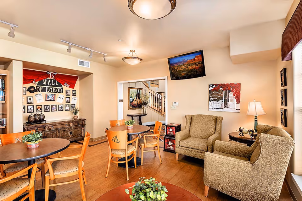 Bright common room with round dining tables, wooden chairs, upholstered armchairs, wall decor and a mounted TV.