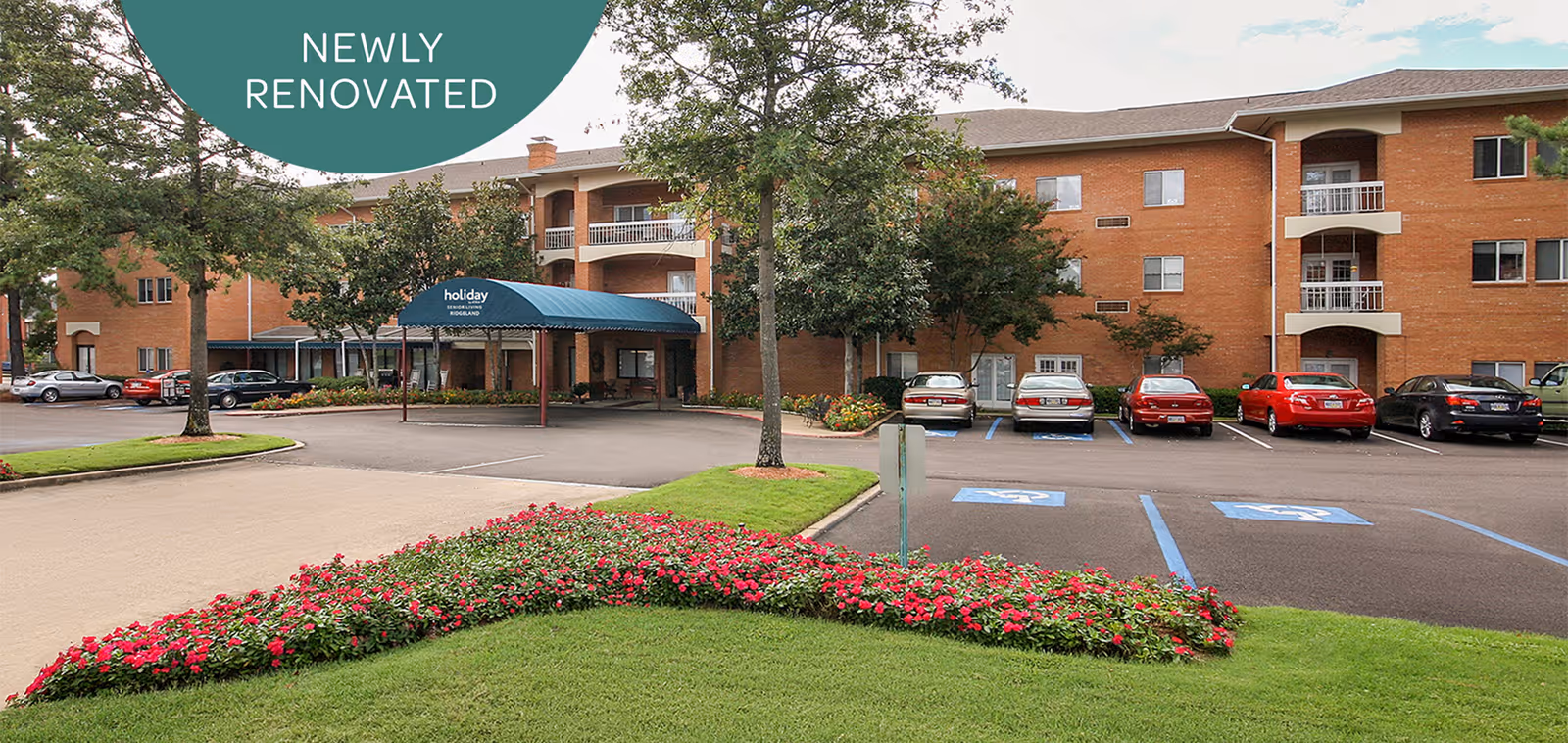 Exterior view of Holiday Chateau Ridgeland, a three-story brick building with balconies and a covered entrance. The parking lot in front has several cars parked, including handicapped spaces. There are trees and a flower bed with red flowers in the foreground. A green banner in the top left corner reads 'Newly Renovated'.