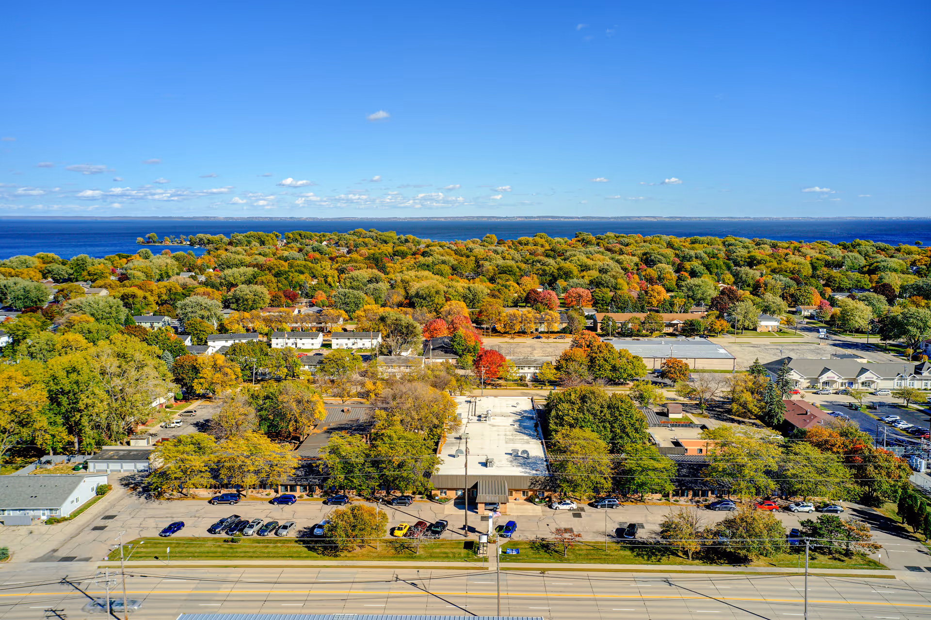 Aerial view of a senior living facility named Edenbrook Oshkosh surrounded by trees with autumn foliage, parking lots with cars, and a large body of water in the background under a clear blue sky.